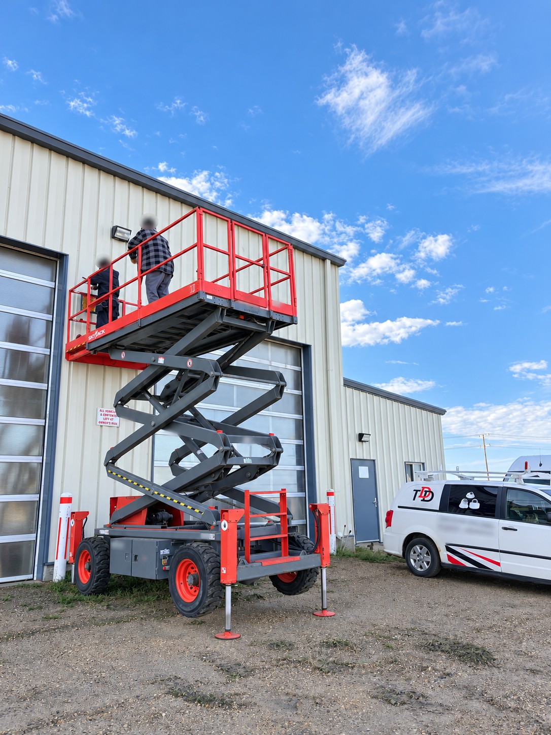 Raised red scissor lift beside a truck wash building during exterior CCTV installation, with identifying details blurred